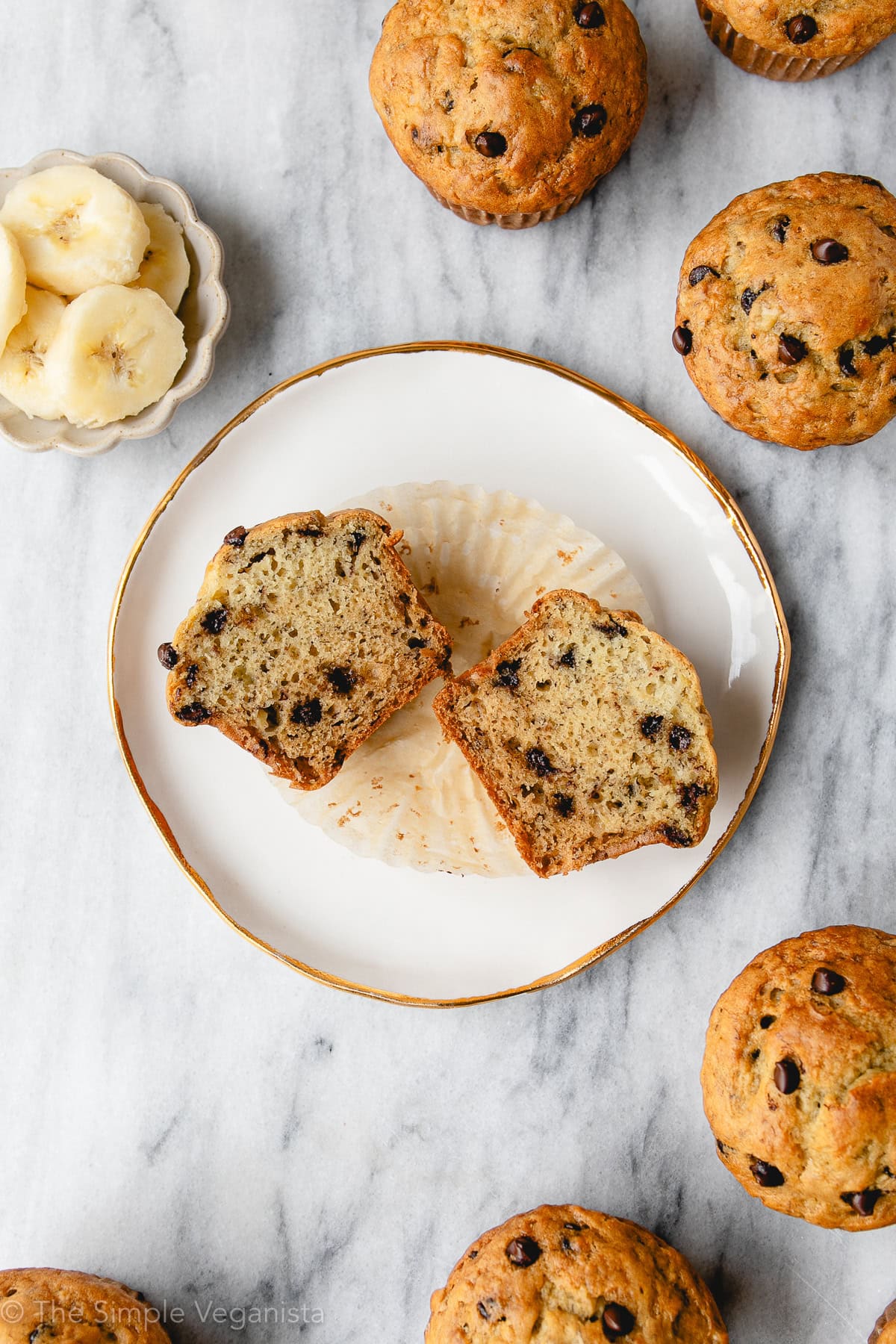 Freshly baked vegan banana chocolate chip muffin split in half facing up on a white plate with items surrounding.