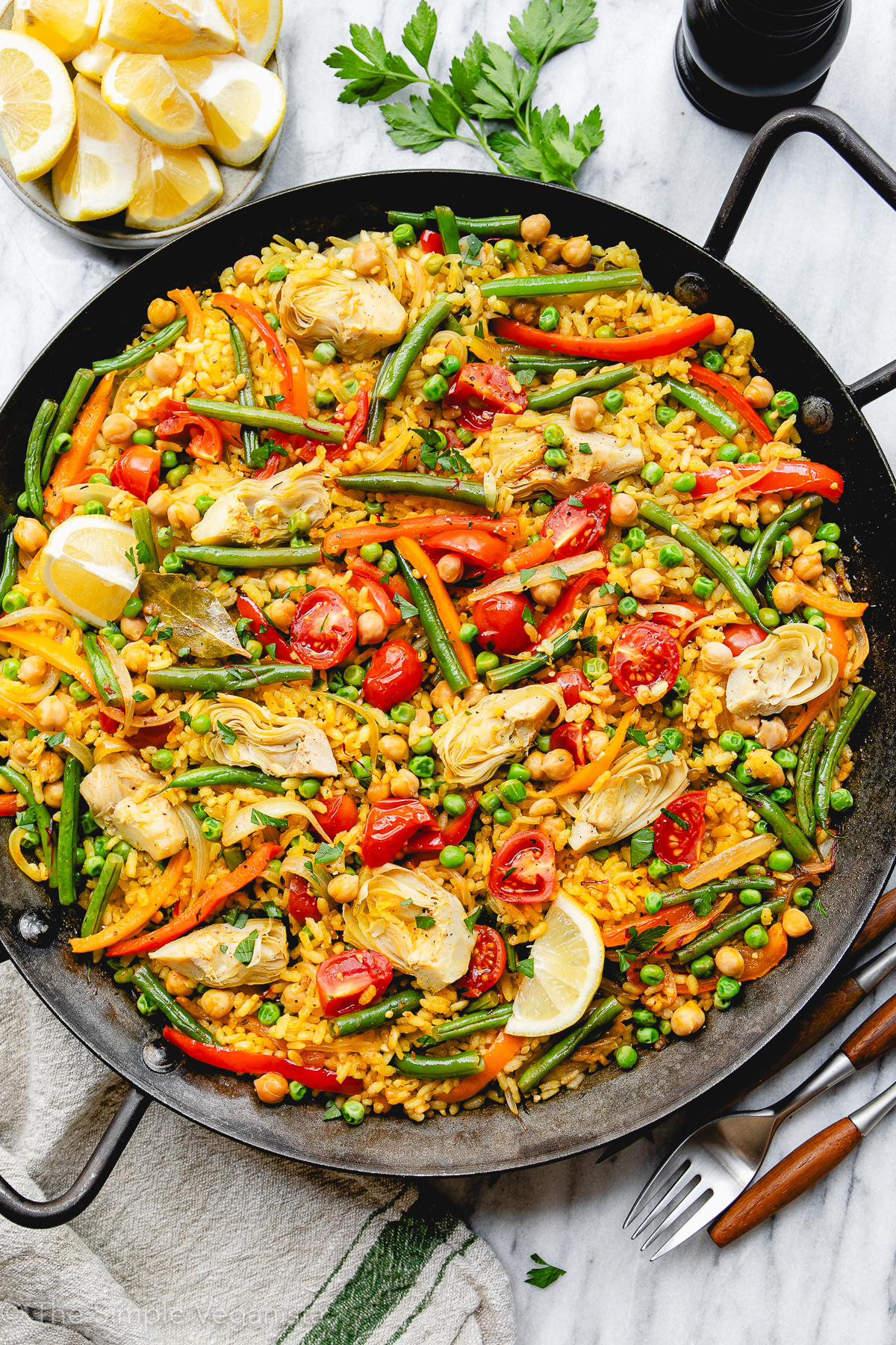 Colorful vegan vegetable paella made with fresh vegetables, saffron rice, and herbs in a large pan with items surrounding.